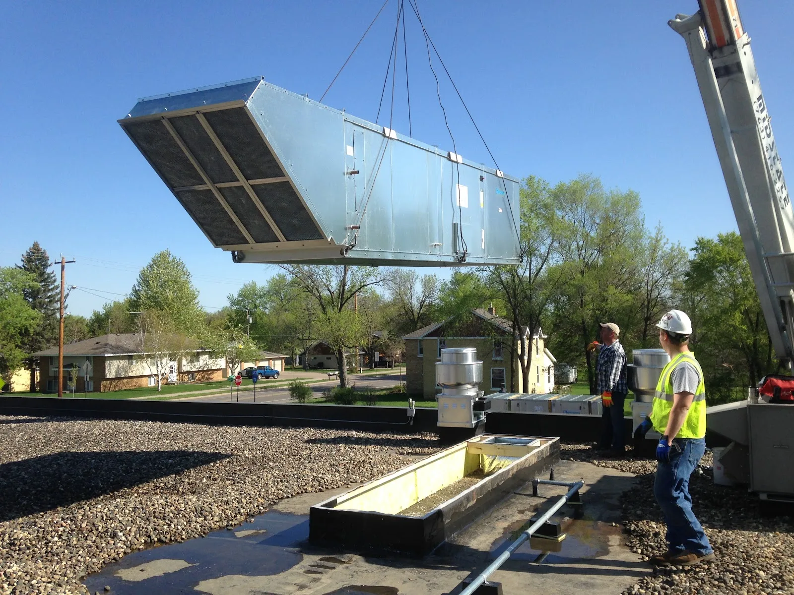 Two workers guiding a large rooftop air handling unit lifted by a crane onto a commercial flat roof